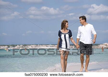 Stock Image - Couple walking on beach holding hands, portrait. Fotosearch