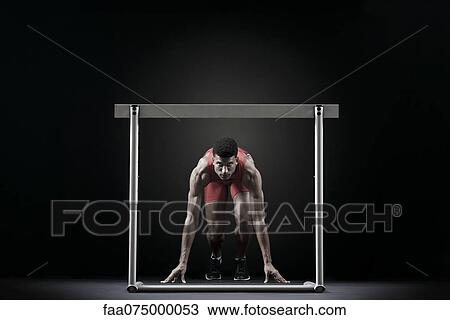 Stock Image - Male athlete at starting position in front of hurdle. Fotosearch