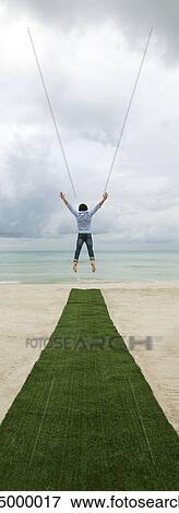 Stock Photo - Man above artificial turf on beach, arms outstretched, rear view. Fotosearch