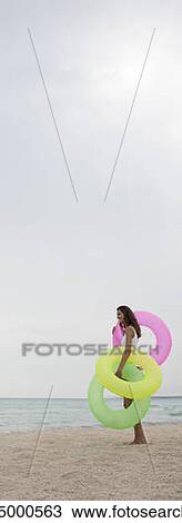 Stock Image - Woman standing on beach, holding inflatable rings, side view. Fotosearch