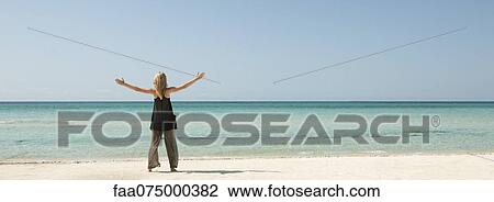 Stock Image - Woman standing on beach with arms outstretched, rear view. Fotosearch