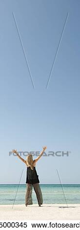 Stock Photography - Woman standing on beach with arms raised in air, rear view. Fotosearch
