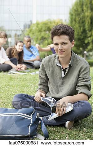 Stock Image - Young man sitting on grass, people in background, portrait. Fotosearch