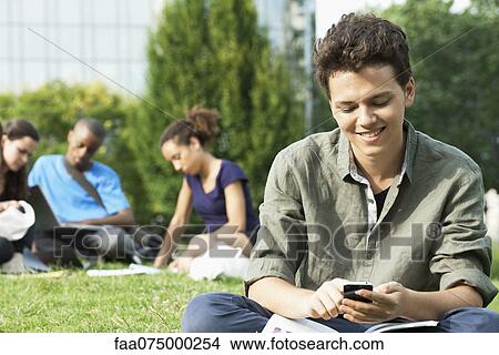 Young man text messaging with cell phone, group of people in background View Large Photo Image Picture - Young man text messaging with cell phone, group of people in background. Fotosearch