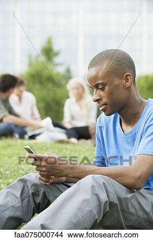 Young man text messaging with cell phone, people in background View Large Photo Image Picture - Young man text messaging with cell phone, people in background. Fotosearch