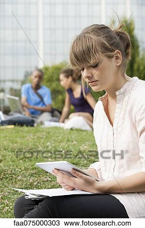 Stock Image - Young woman reading electronic book on grass, people in background. Fotosearch