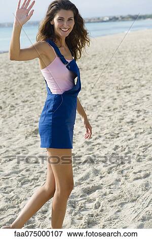 Stock Photo - Young woman walking on beach, waving at camera. Fotosearch