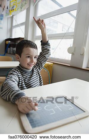 Boy sitting at desk with chalkboard, hand raised View Large Photo Image Stock Image - Boy sitting at desk with chalkboard, hand raised. Fotosearch
