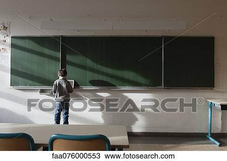 Stock Image - Boy standing in classroom, in front of blackboard, rear view. Fotosearch