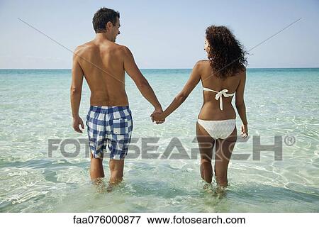 Couple holding hands while wading in sea, rear view View Large Photo Image Stock Photo - Couple holding hands while wading in sea, rear view. Fotosearch