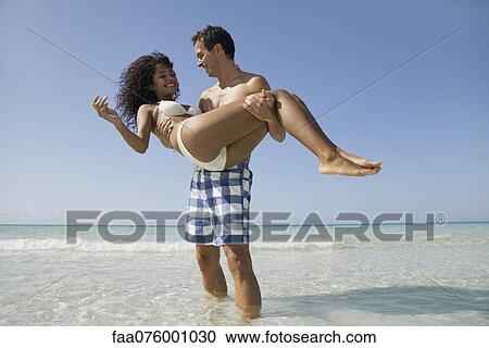 Stock Image - Couple together at the beach, man carrying woman. Fotosearch