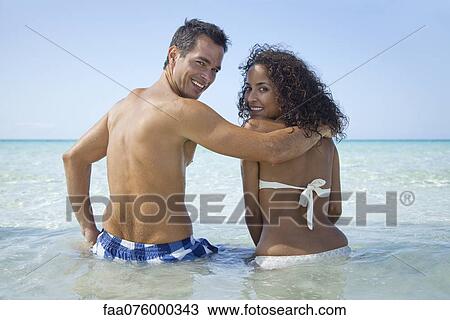 Stock Image - Couple waist deep in water at the beach, smiling over shoulders at camera. Fotosearch