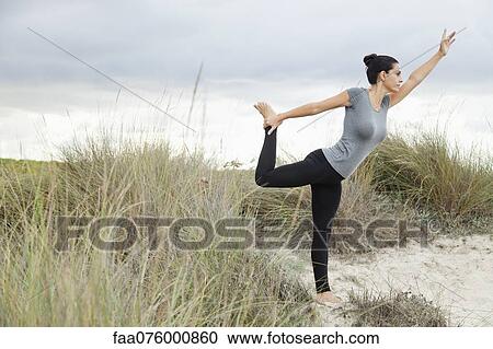 Stock Image - Mature woman in Natarajasana pose on beach, side view. Fotosearch
