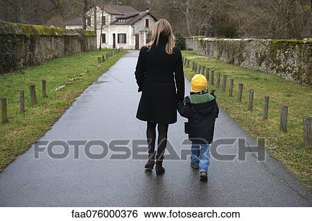 Stock Photograph - Mother and son walking on path in village, holding hands, rear view. Fotosearch