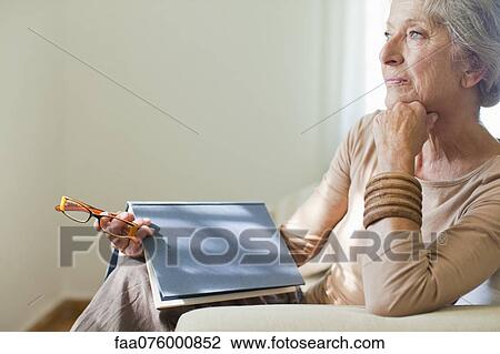 Stock Image - Senior woman sitting on sofa with book. Fotosearch