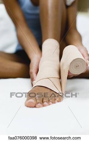 Stock Photo - Woman wrapping bandage around injured ankle, low section. Fotosearch