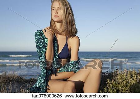 Stock Photograph - Young woman in bikini sitting on beach, portrait. Fotosearch