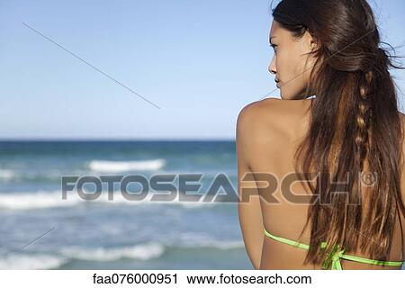 Stock Image - Young woman looking at ocean view, rear view. Fotosearch