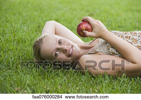 Picture - Young woman lying on grass holding apple. Fotosearch