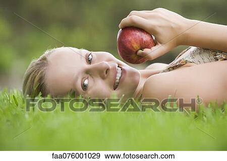 Stock Photo - Young woman lying on grass holding apple. Fotosearch