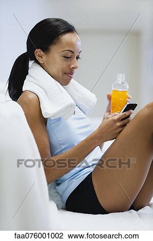 Stock Image - Young woman relaxing with sports drink and text messaging with cell phone. Fotosearch