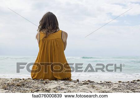 Stock Photo - Young woman sitting on beach looking at view, rear view. Fotosearch