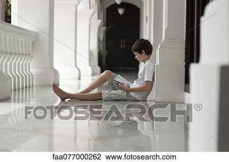 Stock Image - Boy sitting on floor of veranda reading book. Fotosearch