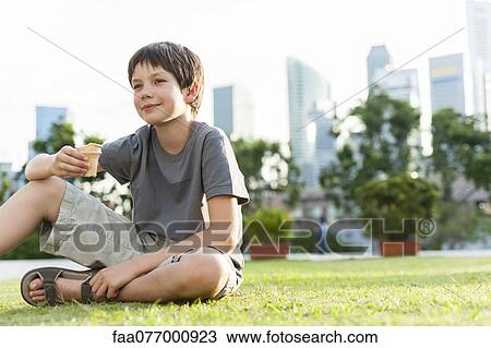 Stock Image - Boy sitting on grass in park, city skyline in background. Fotosearch