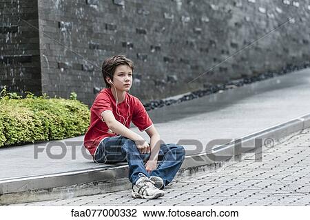 Stock Image - Boy sitting on sidewalk listening to music with earphones. Fotosearch