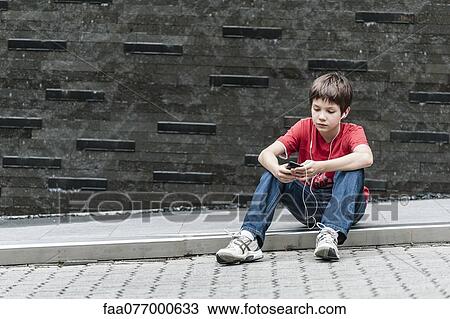 Stock Image - Boy sitting on sidewalk listening to music with earphones. Fotosearch