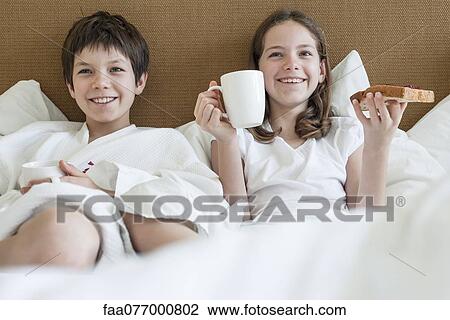 Stock Image - Brother and sister lying in bed eating breakfast. Fotosearch