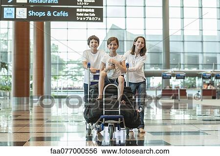 Stock Photograph - Brother and sister pushing younger sister on luggage cart in airport. Fotosearch