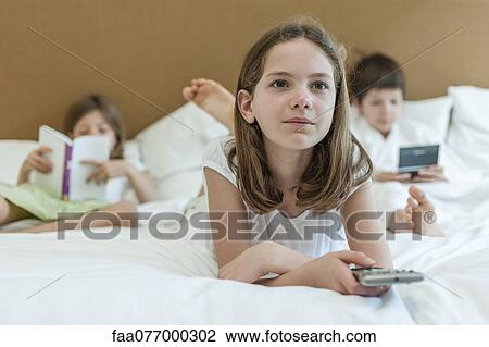 Girl lying on bed watching TV, siblings in background View Large Photo Image Stock Image - Girl lying on bed watching TV, siblings in background. Fotosearch