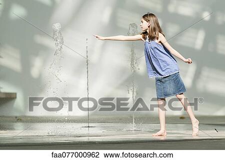Stock Image - Girl reaching for vertical water jet rising from public fountain. Fotosearch