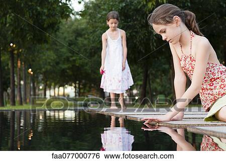 Stock Photograph - Girl sitting by edge of pond touching surface of water, another girl in background. Fotosearch