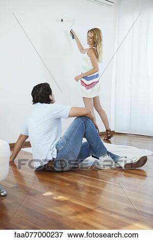 Stock Photo - Man watching woman painting wall using paint roller. Fotosearch
