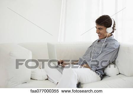Stock Photography - Mature man using laptop computer on sofa wearing headphones. Fotosearch