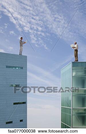 Stock Photo - Men standing on top of high rise buildings, using laptop computers. Fotosearch