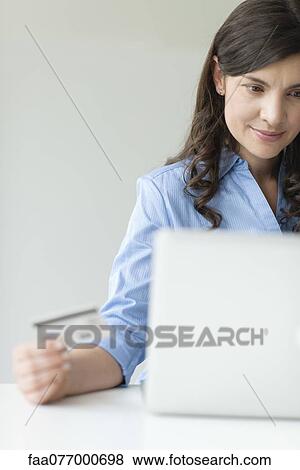 Stock Photo - Mid-adult woman holding credit card while using laptop computer. Fotosearch