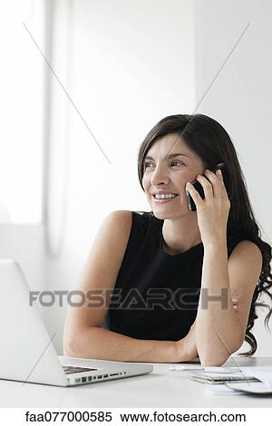 Stock Photography - Mid-adult woman sitting at desk with laptop, talking on cell phone. Fotosearch