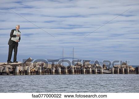 Stock Image - Oversized businessman standing on breakwater, holding briefcase stuffed with money. Fotosearch