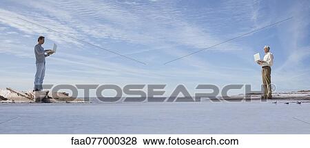 Stock Photo - Oversized men standing on separate shores, using laptop computers. Fotosearch