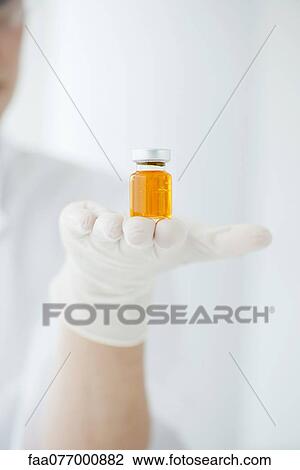 Stock Image - Scientist holding vial containing orange liquid. Fotosearch
