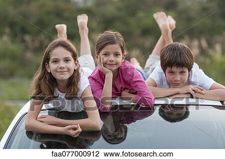 Stock Image - Siblings lying on top of car roof, portrait. Fotosearch
