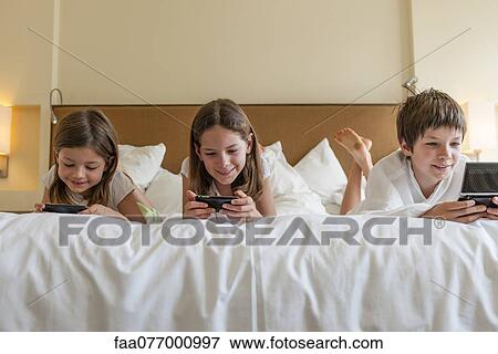 Stock Photo - Siblings playing handheld video games on bed. Fotosearch