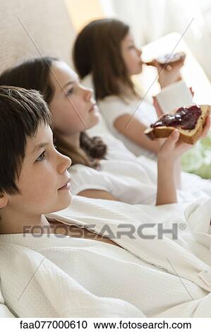 Stock Image - Siblings relaxing on bed, girls eating toasts in background. Fotosearch