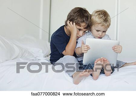 Picture - Young brothers sitting on bed looking at digital tablet. Fotosearch