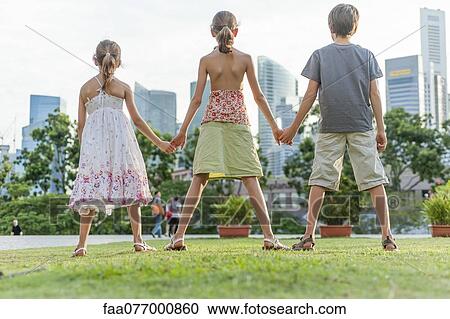 Stock Image - Young siblings standing on grass holding hands, rear view. Fotosearch