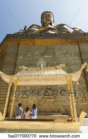 Stock Image - Young tourists standing beneath large statue of buddha. Fotosearch