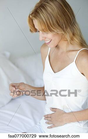 Stock Photo - Young woman holding pregnancy test with hand on abdomen, smiling. Fotosearch